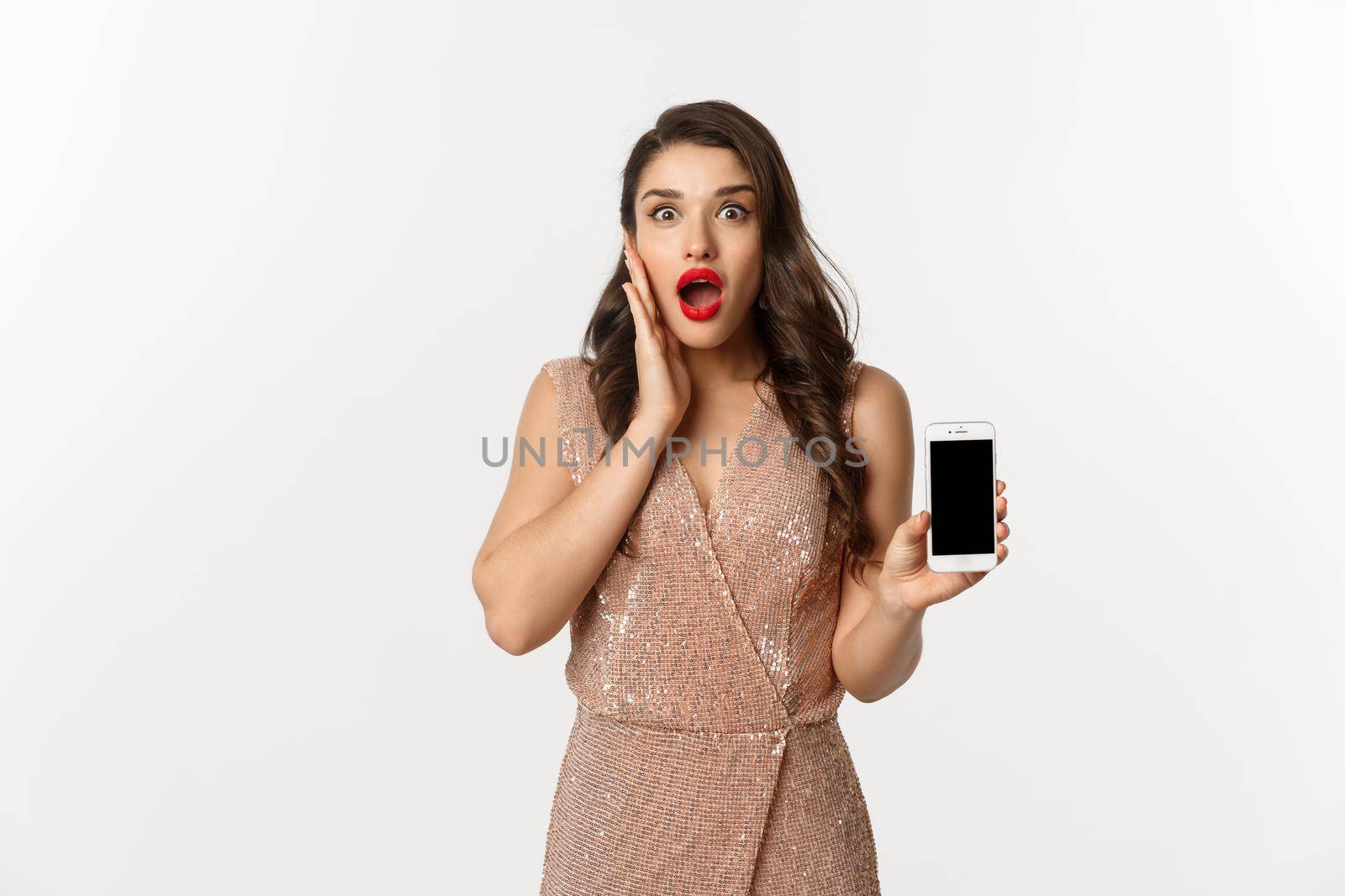 Online shopping. Elegant woman in luxury dress showing smartphone screen, looking amazed at camera, white background.