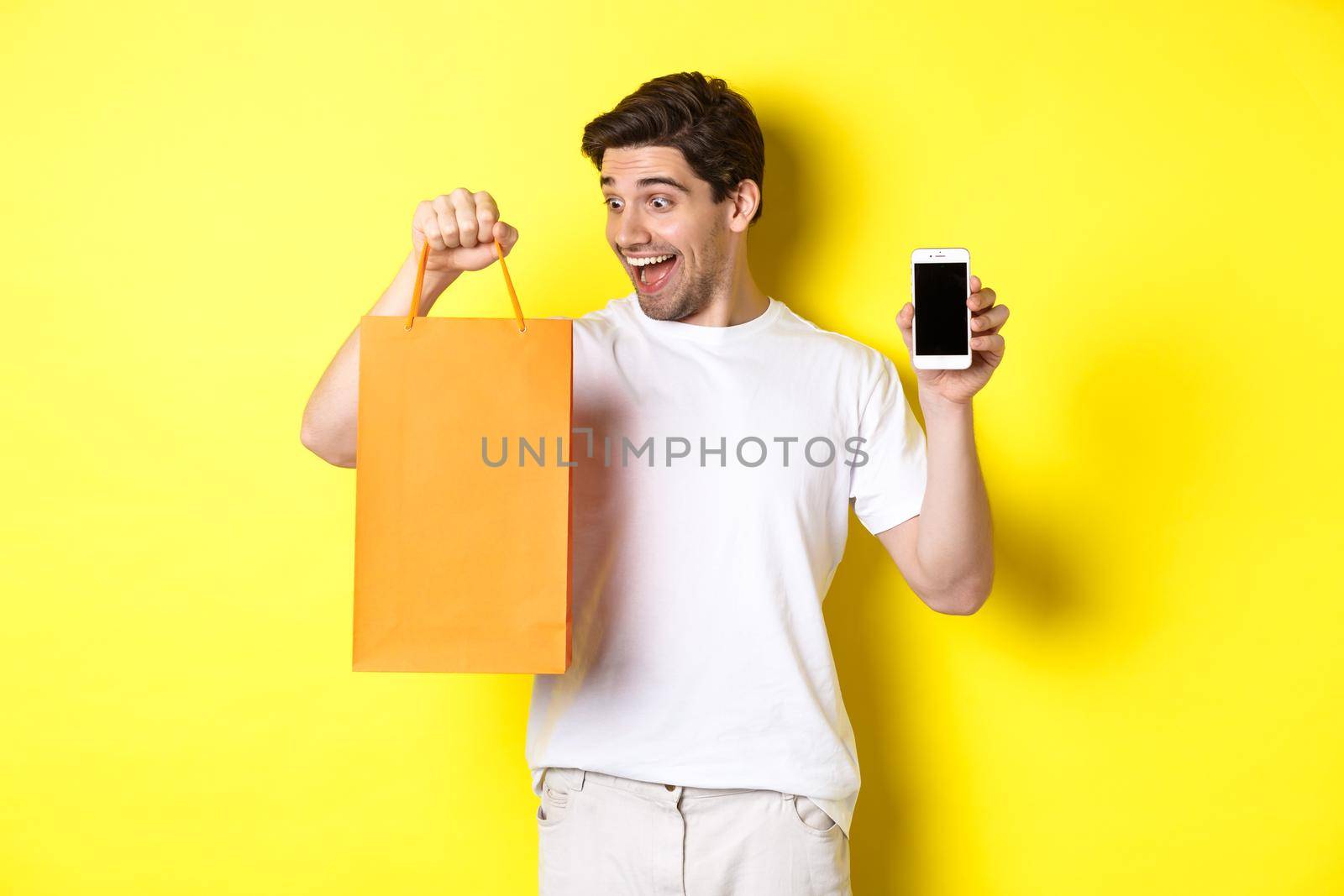 Concept of discounts, online banking and cashback. Happy guy buy something in store and looking at shopping bag, showing mobile phone screen, yellow background.