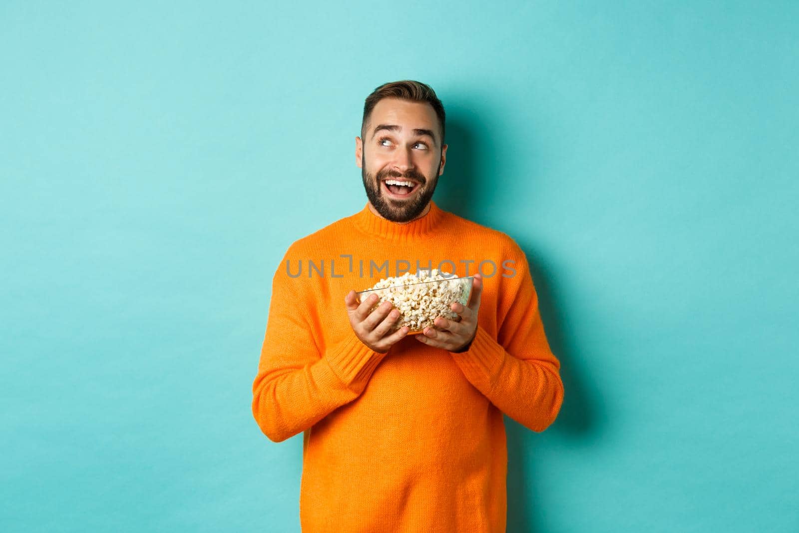 Handsome young man in orange sweater, looking thoughtful at upper left corner, holding popcorn, picking movie, blue background by Benzoix