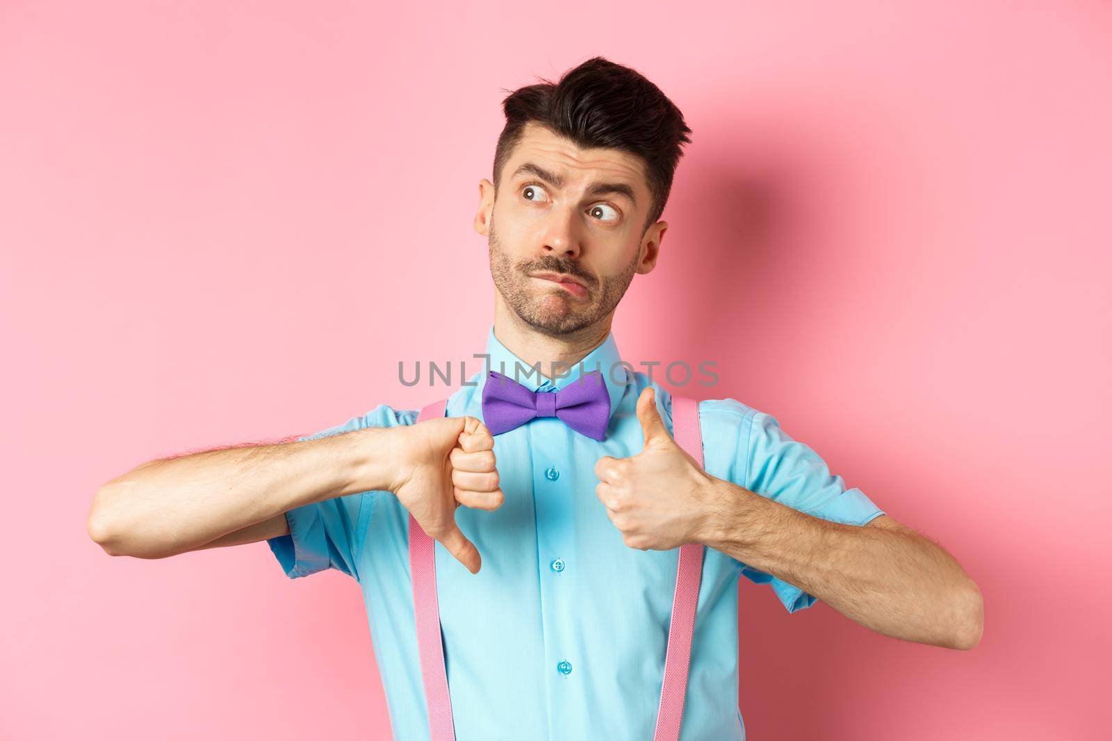 Confused guy looking pensive while judging, showing thumbs up and down, standing indecisive and unsure against pink background.