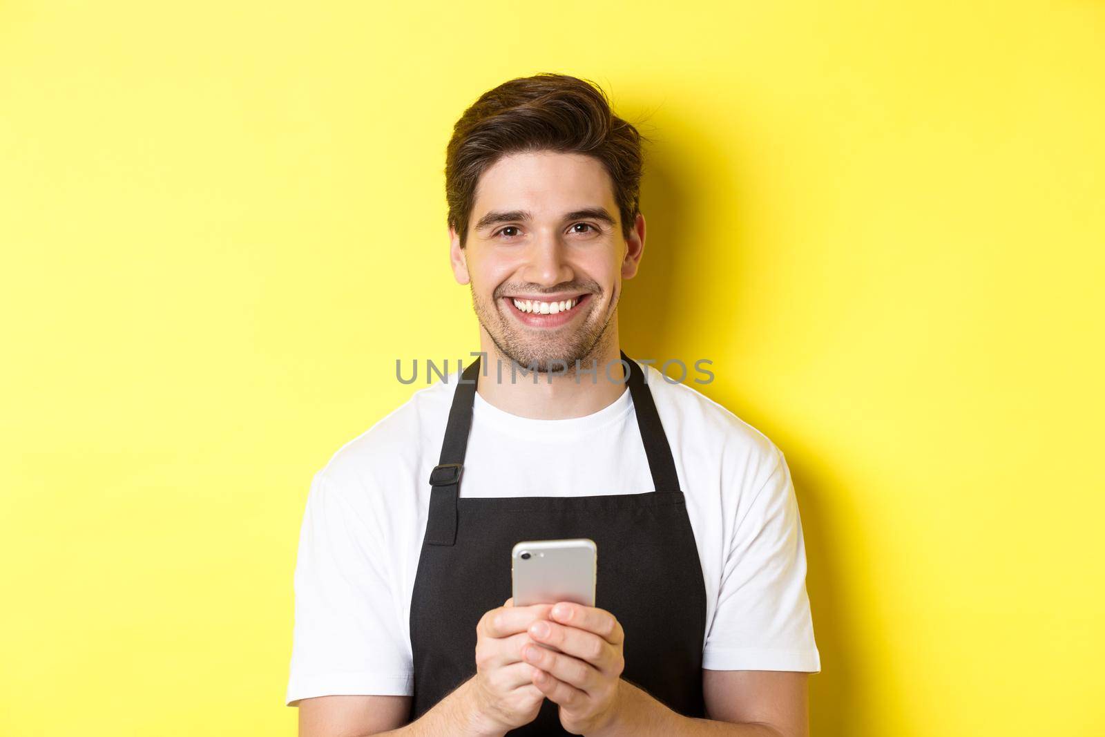 Close-up of handsome barista sending message on mobile phone, smiling happy, standing over yellow background.