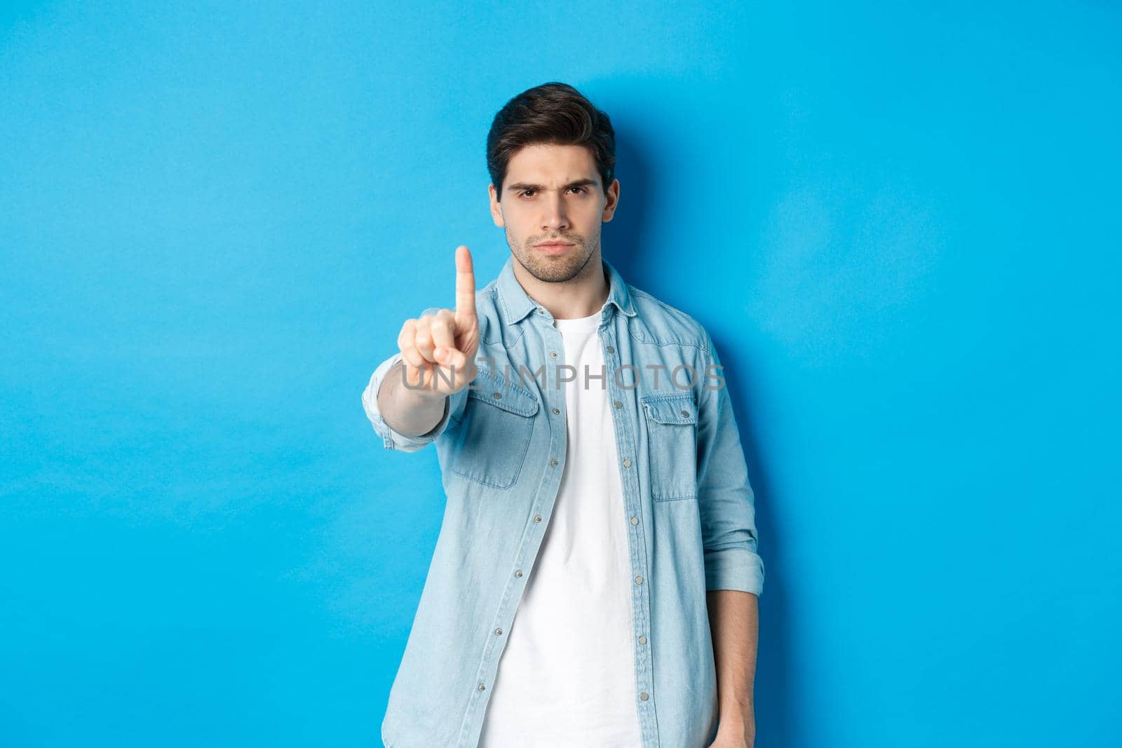 Angry 25 years old man shaking finger in disapproval sign, frowning disappointed, forbid something bad, telling no, standing against blue background.