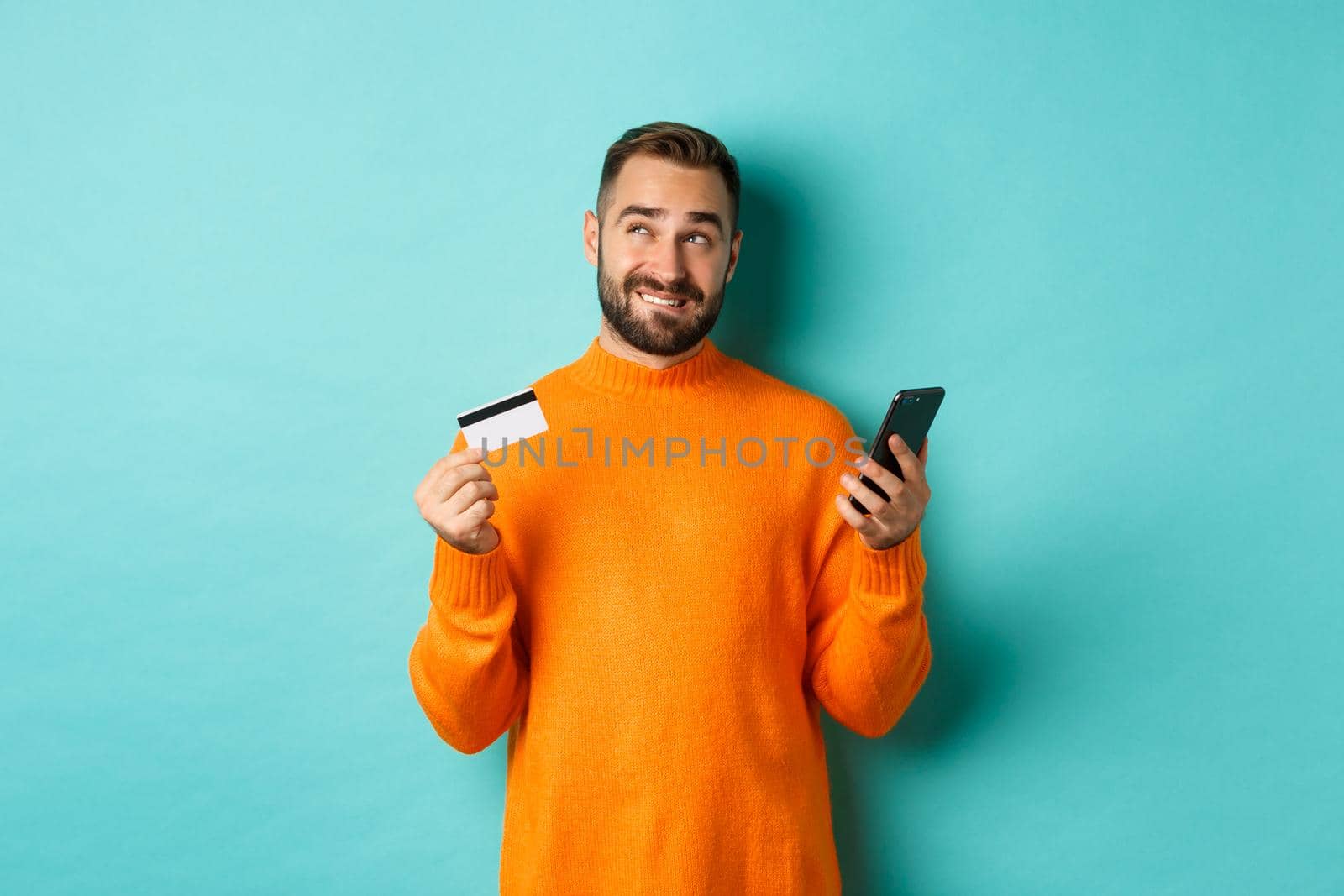 Online shopping. Thoughtful man holding credit card and mobile phone, thinking of purchase, standing over light blue background by Benzoix