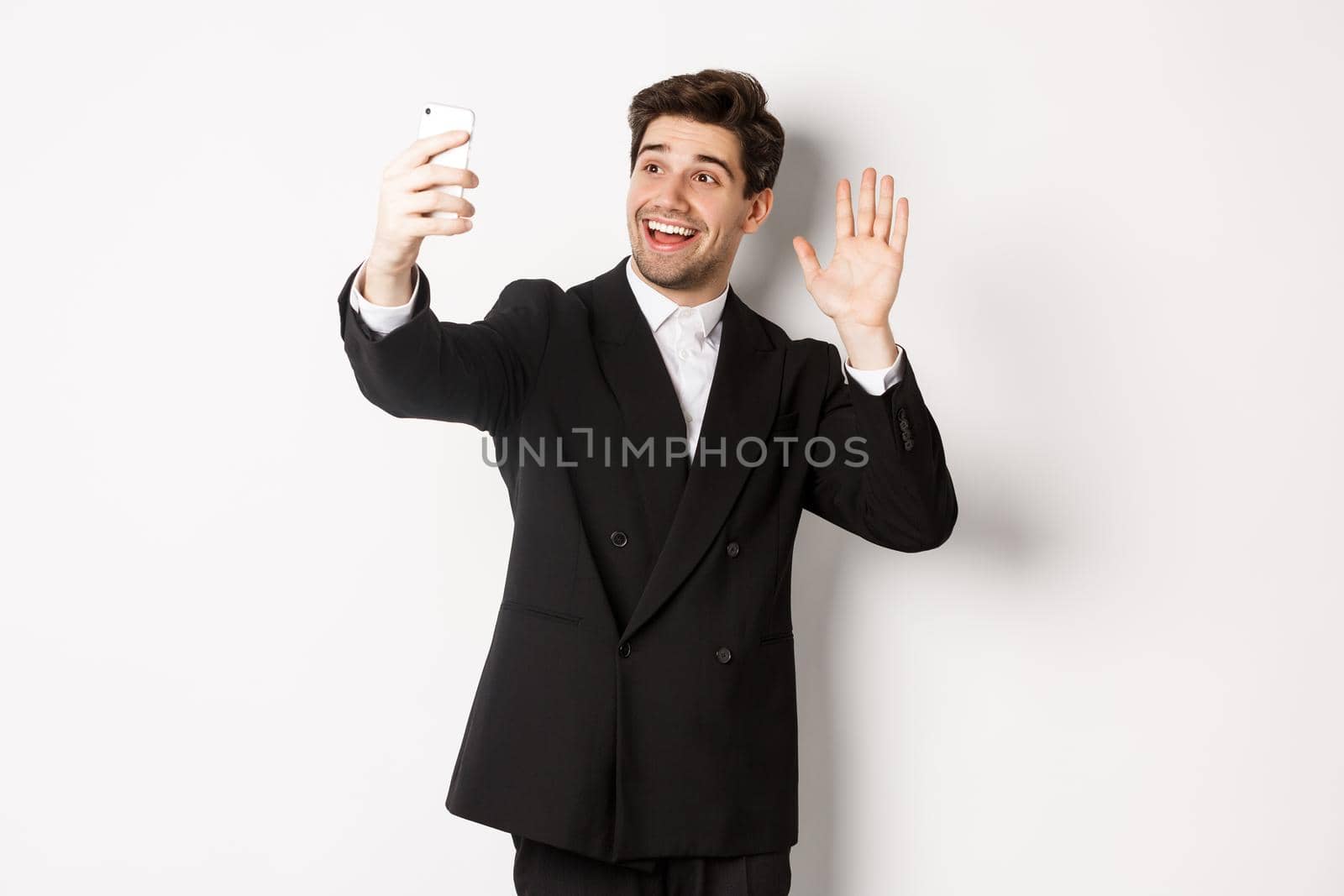 Image of handsome man in suit, having video call and waving hand at smartphone camera, recording video, greeting someone, standing against white background by Benzoix