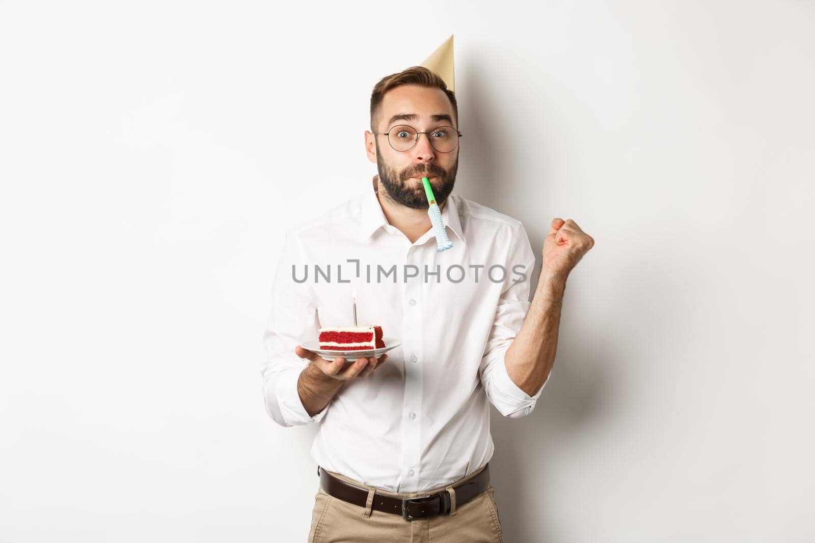 Holidays and celebration. Cheerful man enjoying birthday, blowing party whistle and holding bday cake, white background.