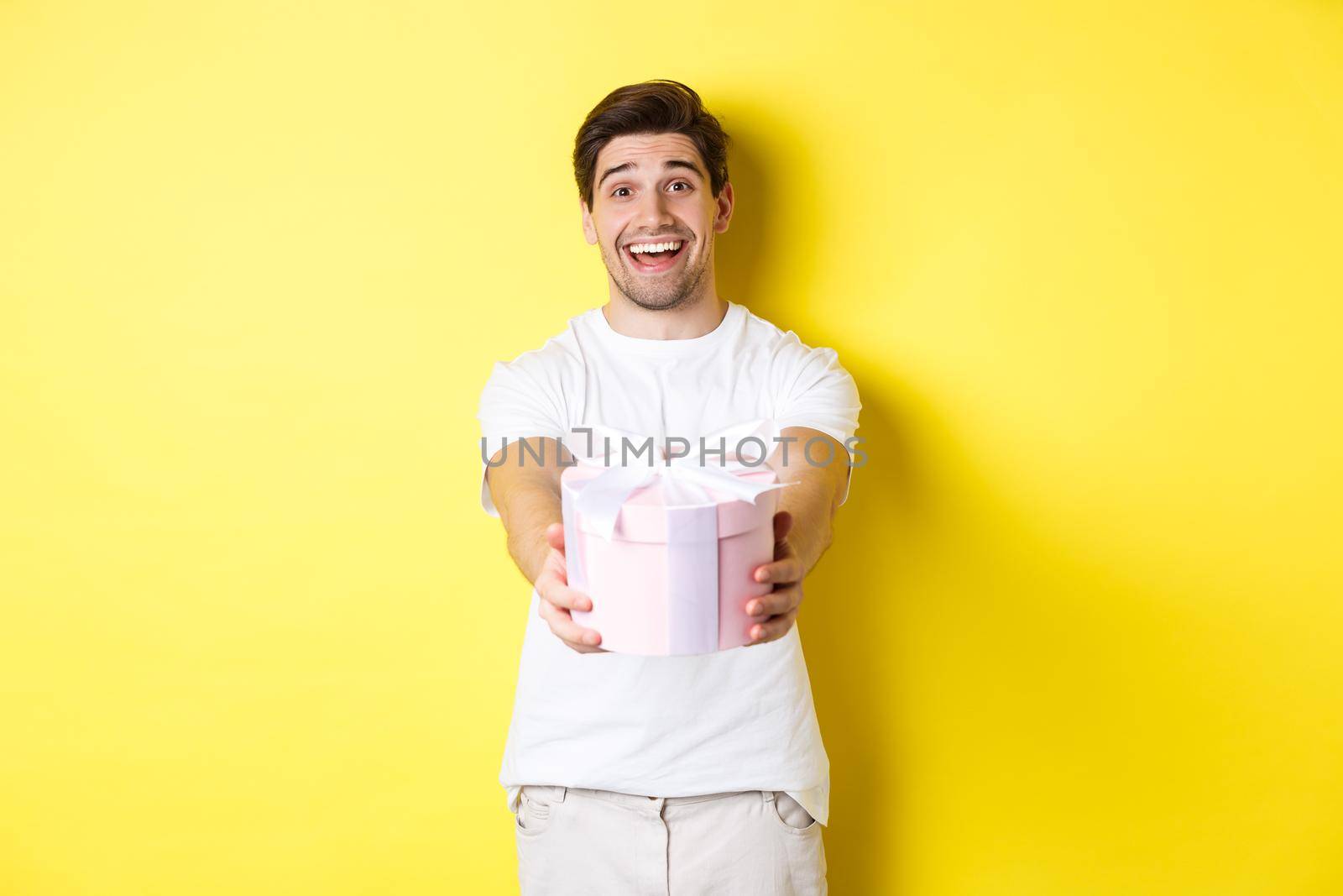 Concept of holidays and celebration. Smiling man giving you gift, congratulating, standing over yellow background with present.