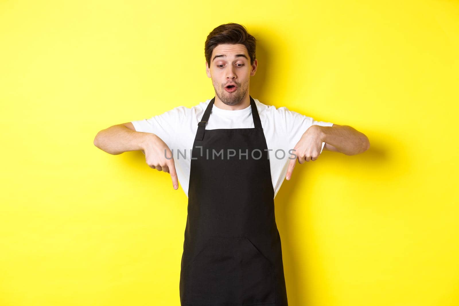 Surprised male barista in black apron checking out promo offer, pointing fingers down at logo banner, standing over yellow background.