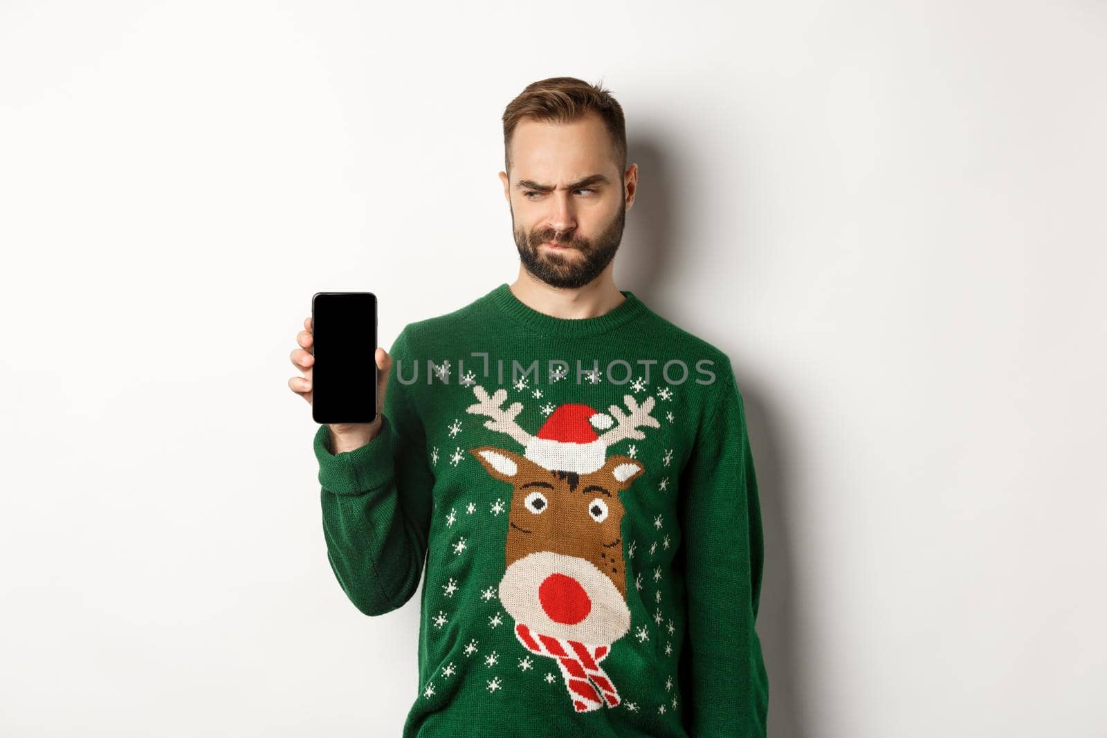 New Year, holidays and celebration. Skeptical guy looking doubtful at screen while showing it to you, demonstrating mobile app, standing over white background.