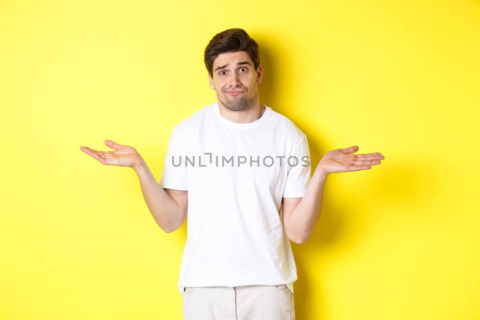 Clueless man in white t-shirt, shrugging and looking puzzled, dont know anything, standing over yellow background.