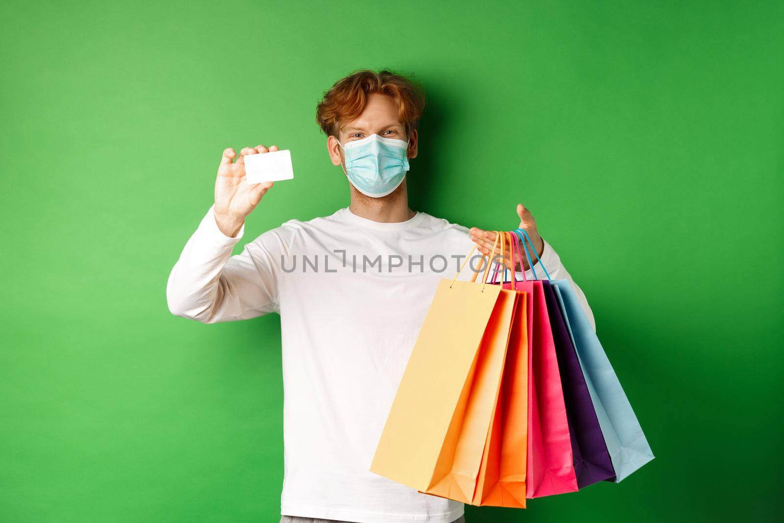 Handsome young man in medical mask, showing plastic credit card and shopping bags with items purchased with discount, green background.