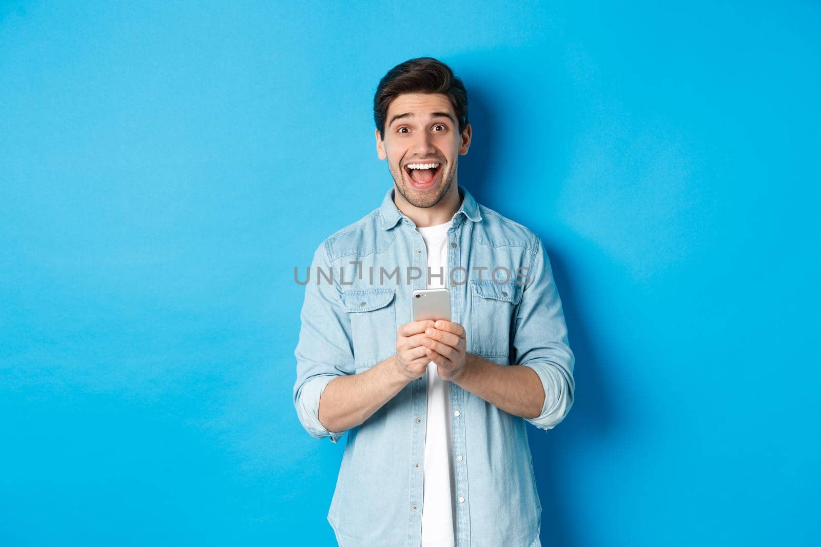 Surprised and happy man winning something online, holding smartphone and rejoicing, standing against blue background.