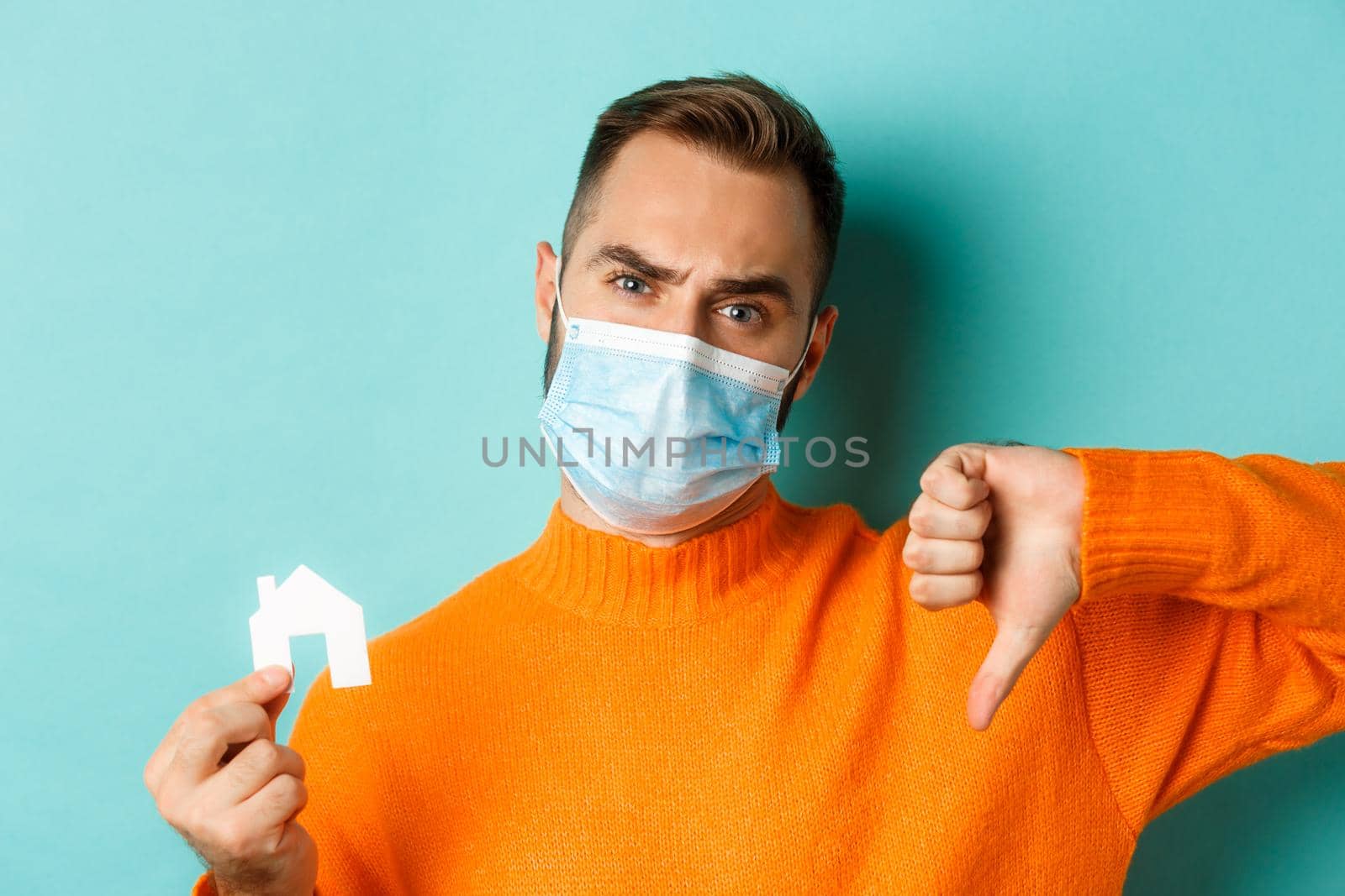 Real estate and coronavirus pandemic concept. Close-up of disappointed man in face mask, showing small paper house and thumb-down, turquoise background.