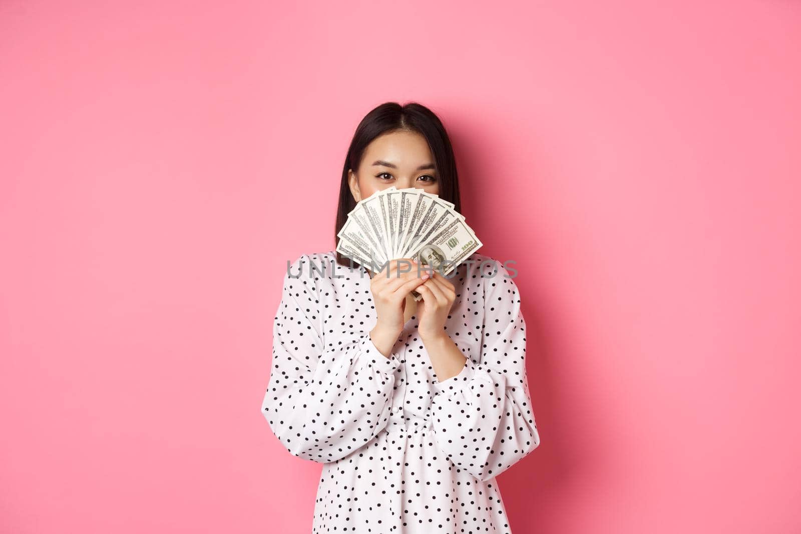 Shopping concept. Cute asian woman hiding face behind money dollars, peeking at camera, standing over pink background by Benzoix
