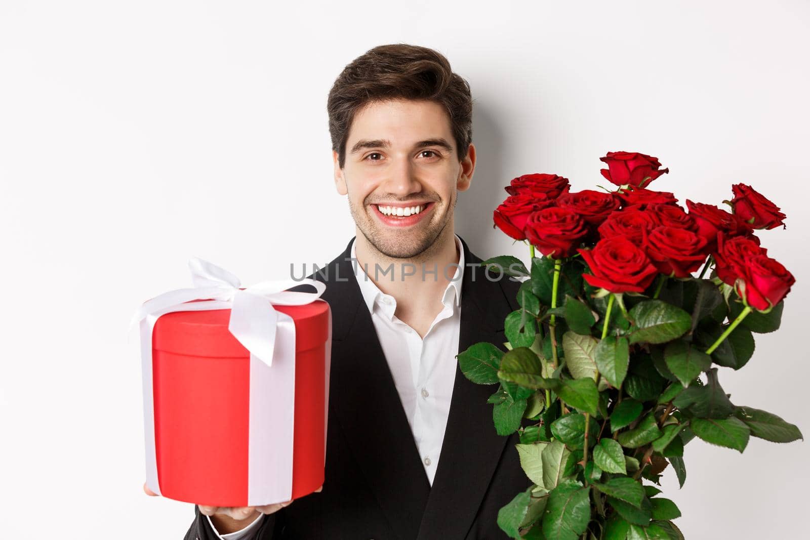 Close-up of handsome bearded man in suit, holding present and bouquet of red roses, smiling at camera, standing against white background.