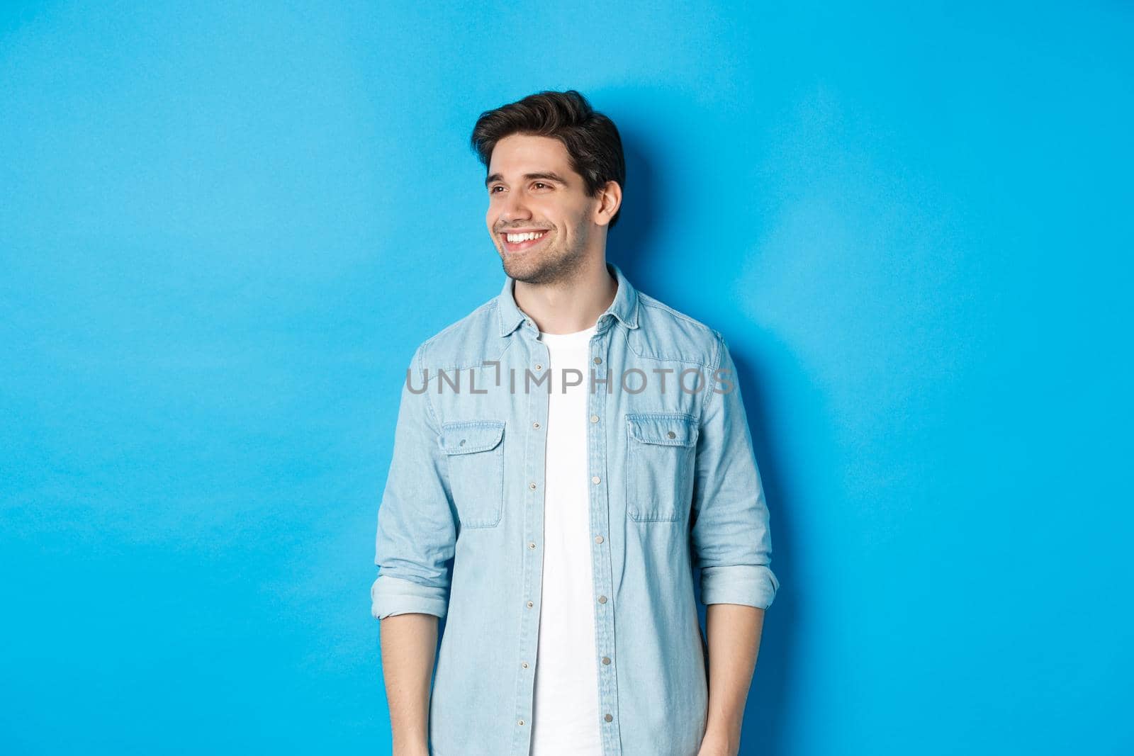 Handsome smiling adult man in casual outfit, smiling and looking left at promo offer, standing against blue background.