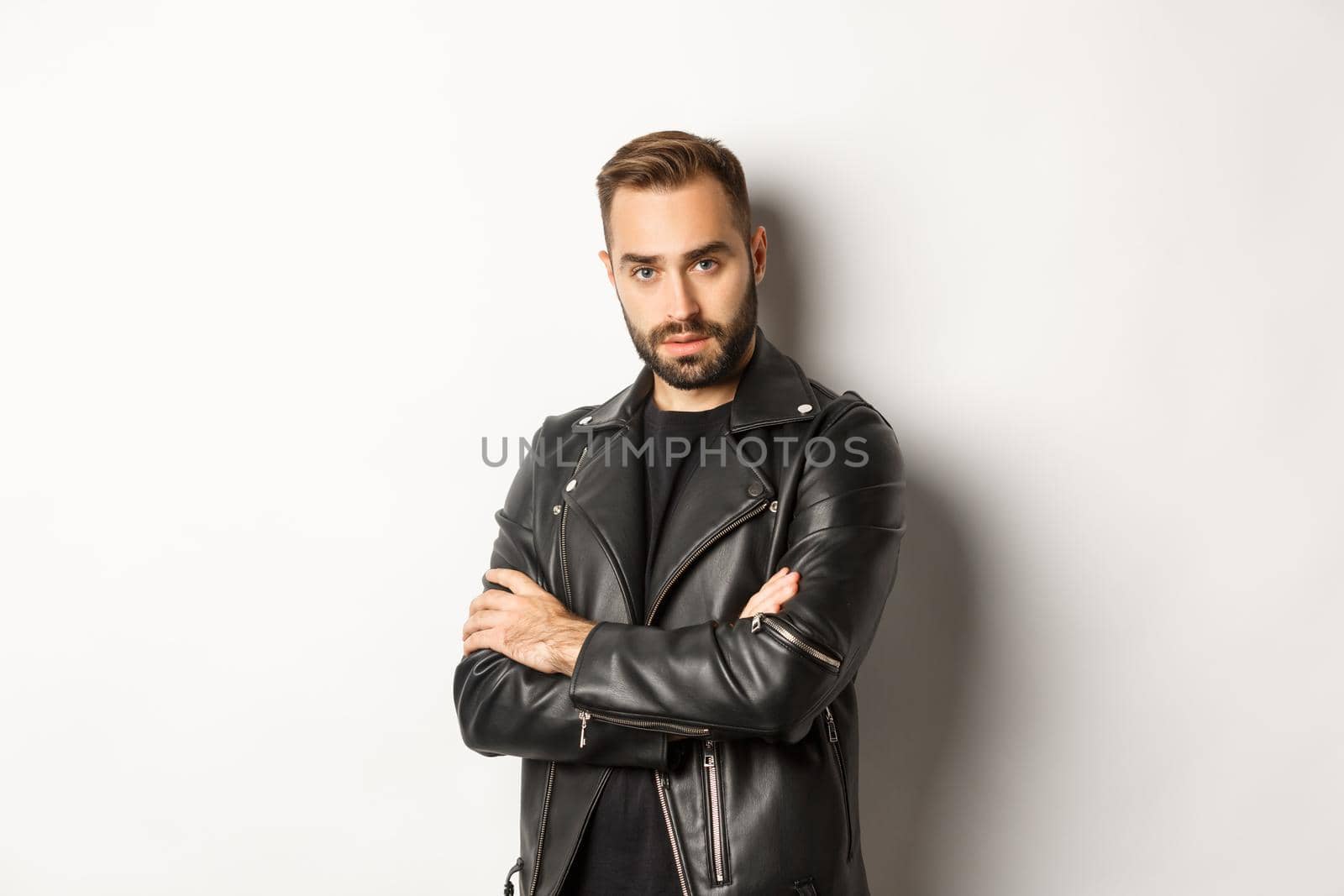 Attractive macho man with beard, wearing leather jacket smiling, looking confident with hands crossed on chest, white background.