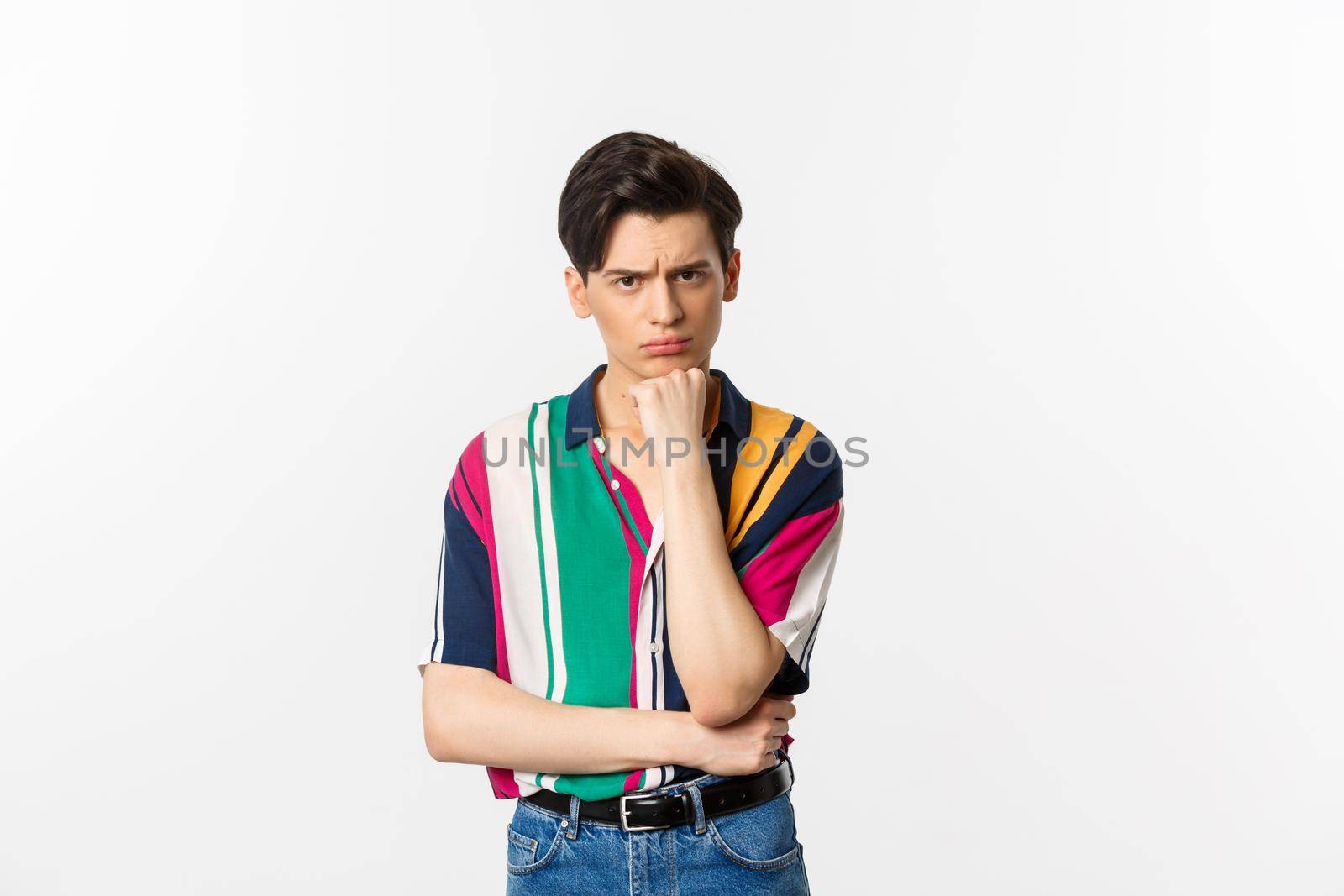 Image of sad gay man looking at camera, sulking displeased and upset, leaning on hand, standing over white background.