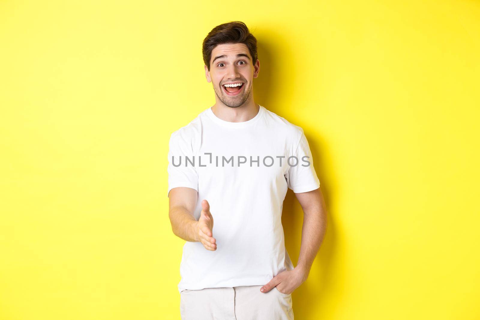 Friendly man greeting you with handshake, smiling amused, saying hello, standing over yellow background.