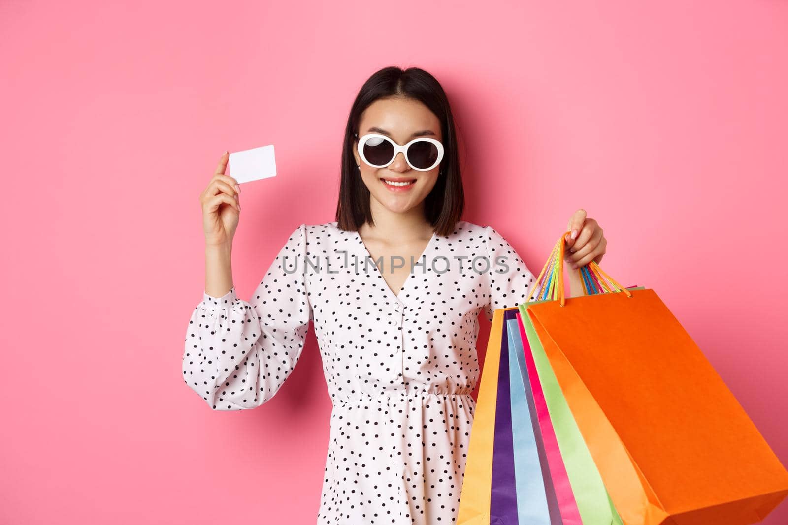 Beautiful asian woman in sunglasses going shopping, holding bags and showing credit card, standing over pink background by Benzoix