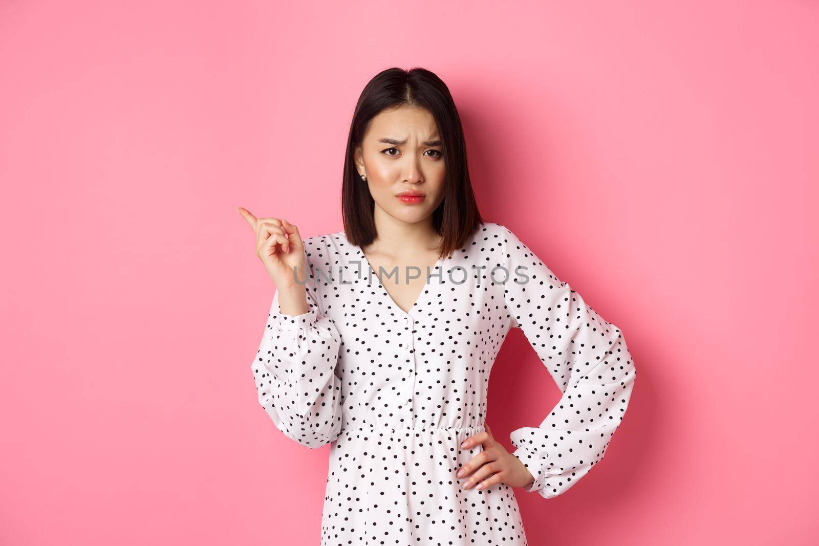 Disappointed asian girlfriend complaining, staring at camera and whining, pointing at upper left corner copy space, standing in dress over pink background by Benzoix
