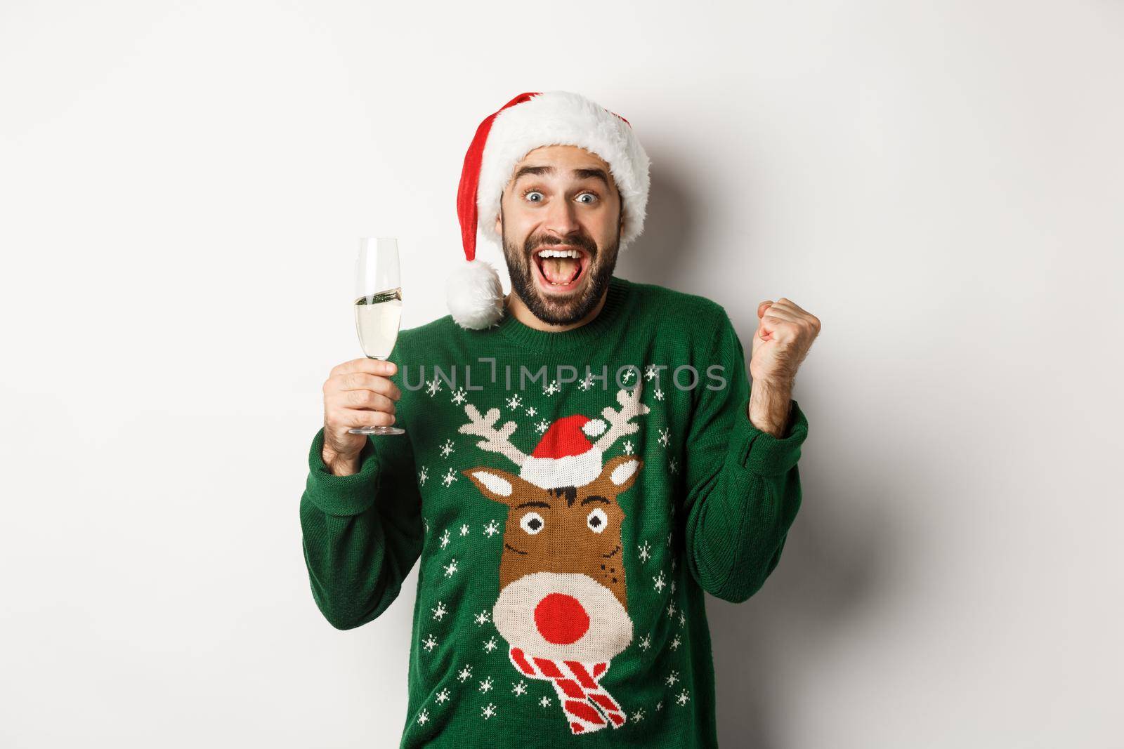 Christmas party and holidays concept. Excited man in Santa hat celebrating New Year, drinking champagne and rejoicing, standing over white background.