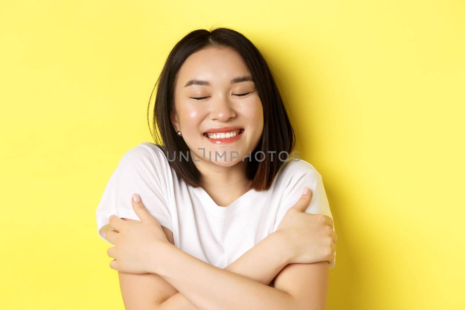 Close up of romantic asian girl hugging herself and dreaming, close eyes and smile while imaging something tender, standing over yellow background.