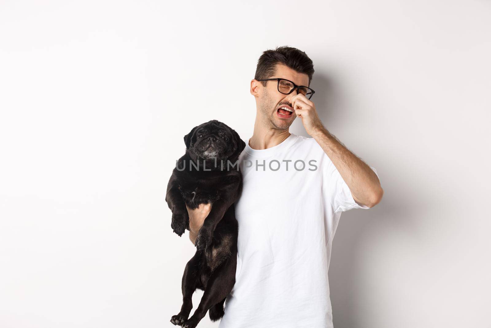 Dog owner squinting and shutting nose from disgusting reek, holding pug with bed smell, standing over white background.