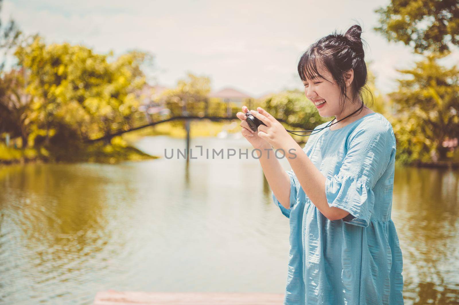 Asian woman in blue dress in public park carrying digital mirrorless camera and taking photo without facial mask in happy mood. People lifestyle and leisure concept. Outdoor travel and Nature theme.