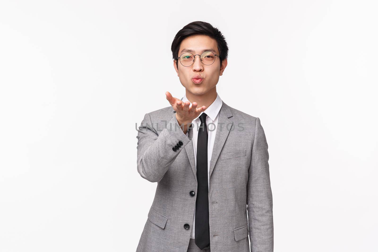 Waist-up portrait of romantic cute, handsome asian man in grey suit, sending air kiss at camera, going to office, saying buy girlfriend staying home, standing white background.