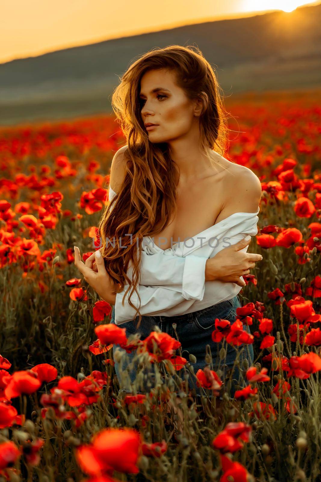 Beautiful young long haired woman in a poppy field at sunset with flying hair. Dressed in a white shirt and denim skirt. by Matiunina