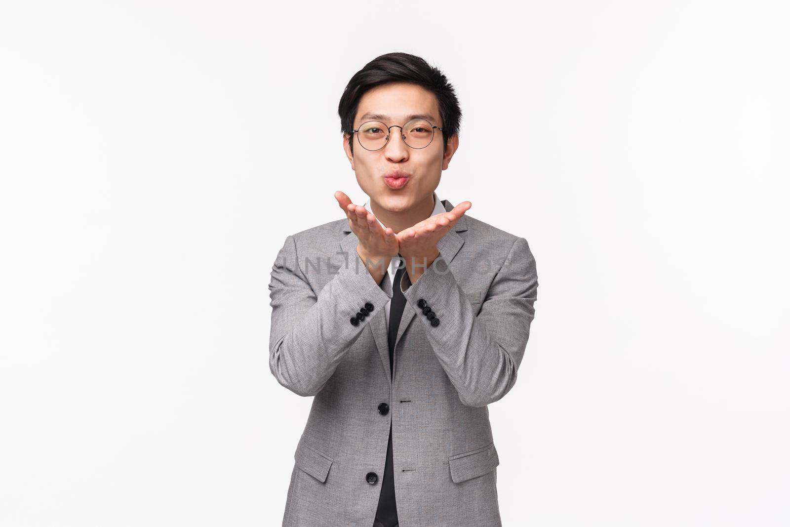 Waist-up portrait of silly handsome, romantic asian boyfriend, businessman in grey suit, holding hands near folded lips to send air kiss at camera, smiling cute, standing white background.