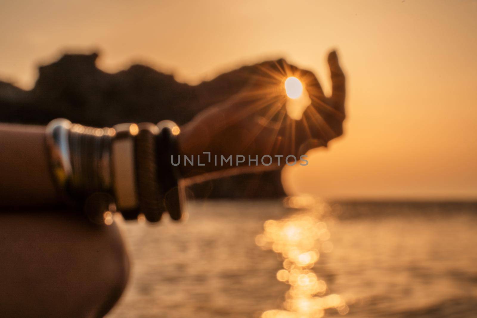 In blur closeup the hand of a young woman in bracelets. Practicing yoga on the beach with sunset. Keeps fingers connected, the sun shines through them. The concept of a healthy lifestyle, harmony