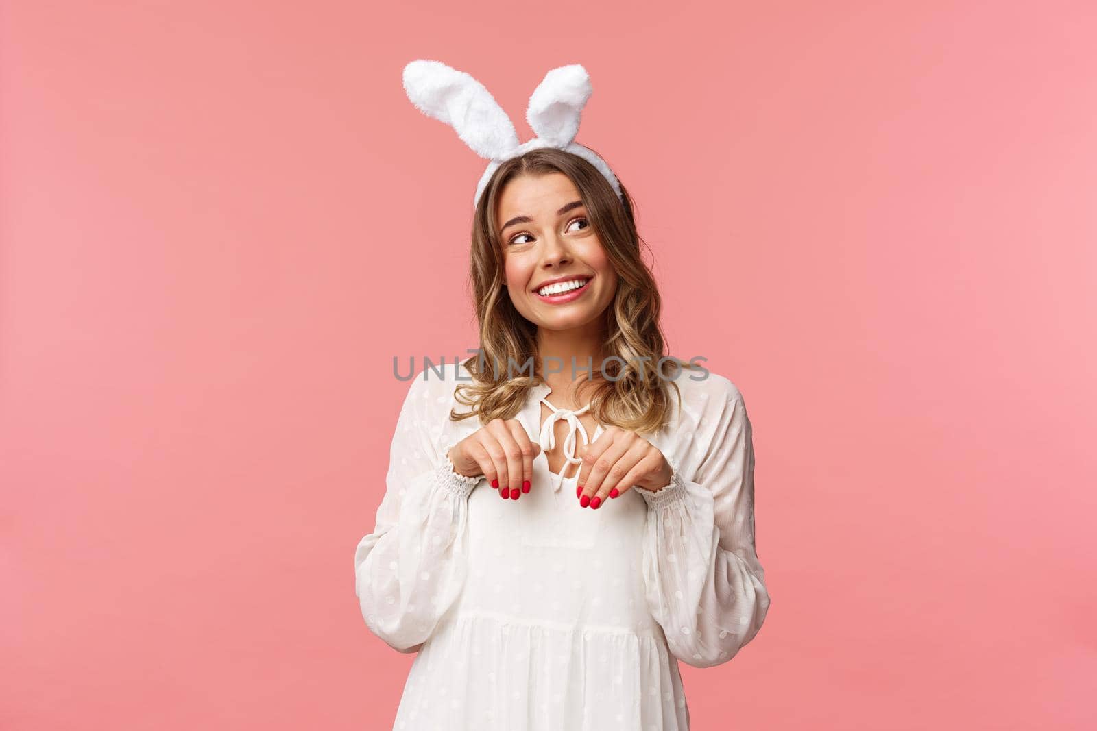 Holidays, spring and party concept. Cute romantic young blond girl imitating bunny, wear white dress lovely rabbit ears, make hand-paws near chest and look dreamy up with daydreaming smile.