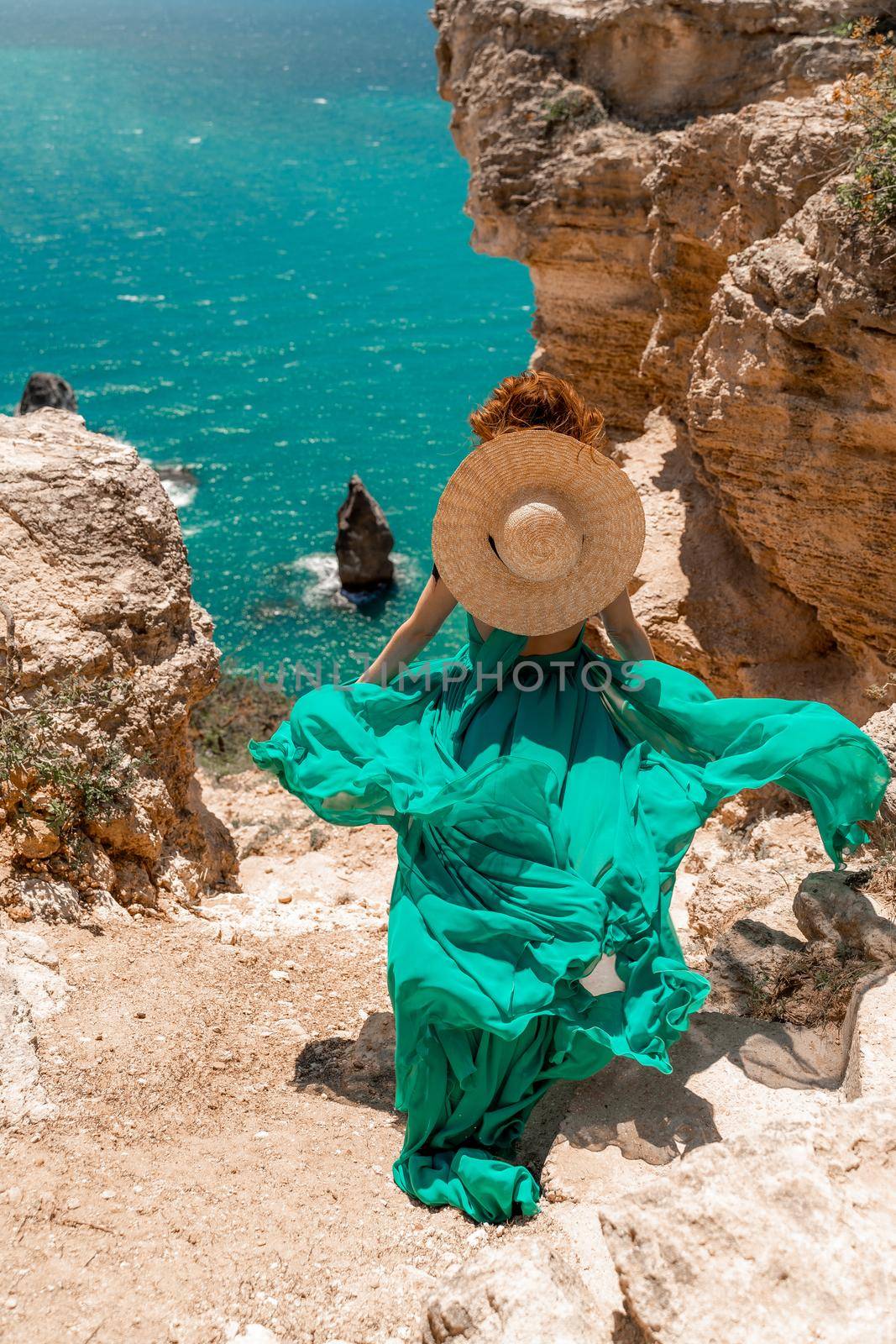 A girl with loose hair in a long mint dress and a straw hat descends the stairs between the yellow rocks overlooking the sea. A strong wind develops the dress. A rock can be seen in the sea. by Matiunina