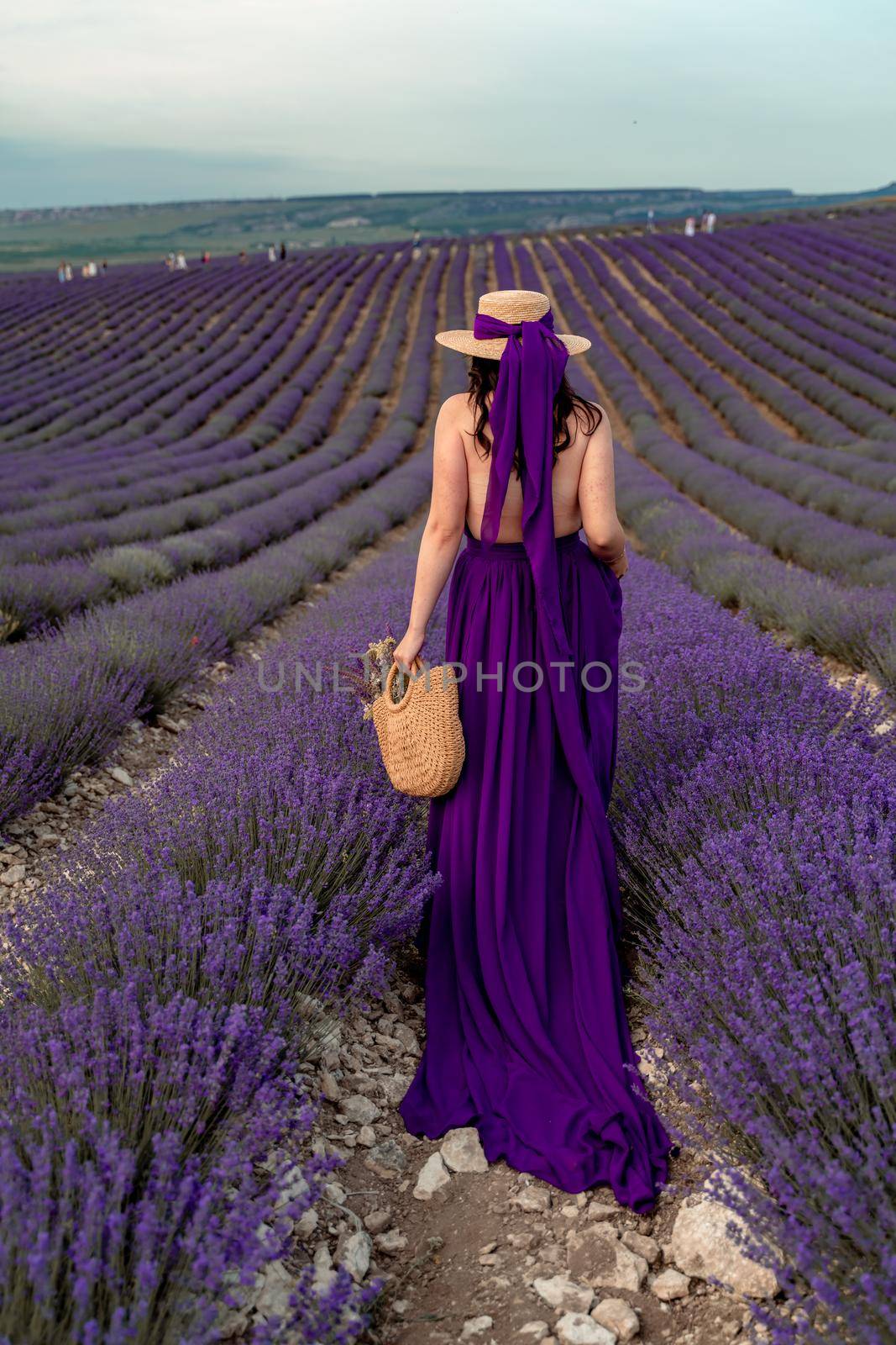 A young beautiful girl in a purple flying dress stands on a blooming lavender field. Rear view. The model has a straw hat by Matiunina