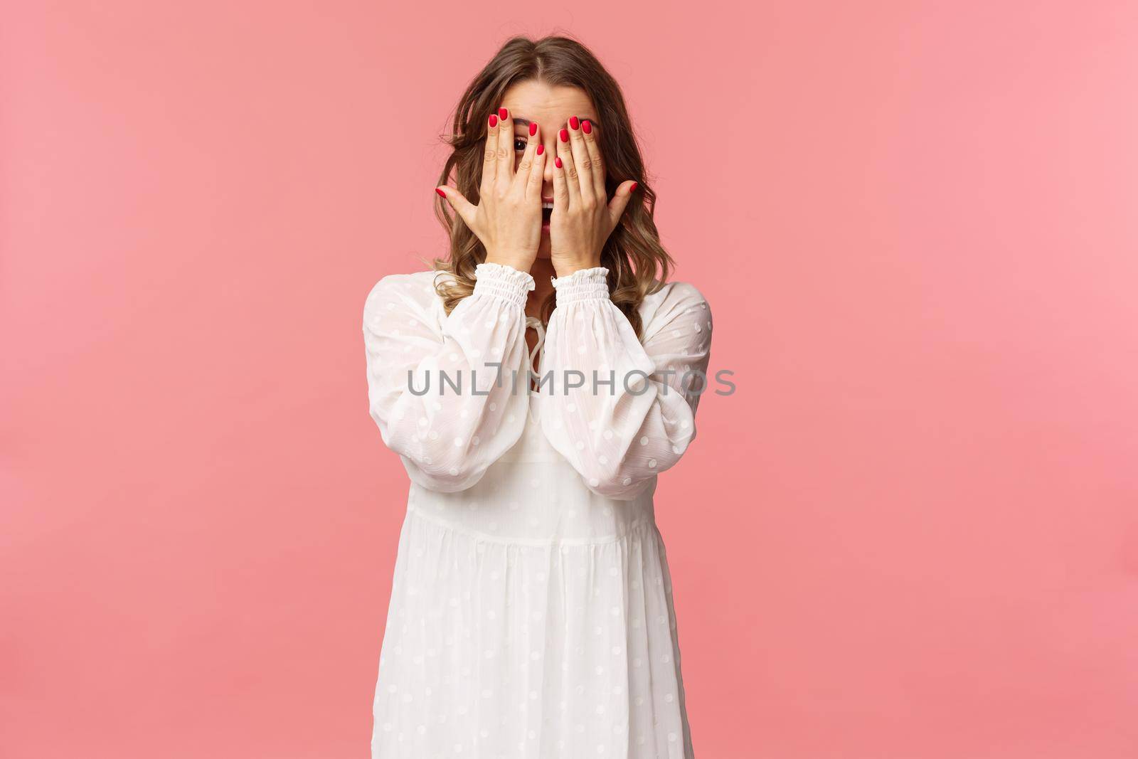 Portrait of silly cute girl with blond short hair, wear white dress, hiding face behind hands, promise to wait for signal but peeking through fingers, cant resist temptation, stand pink background.