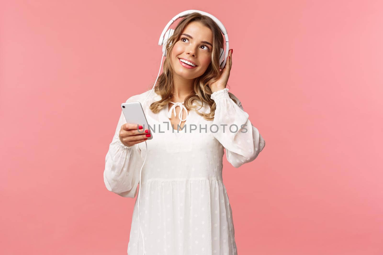 Portrait of tender, beautiful blond caucasian girl in white dress, daydreaming while listening favorite romantic track, wear headphones, hold mobile phone look up thoughtful, enjoy favorite track by Benzoix