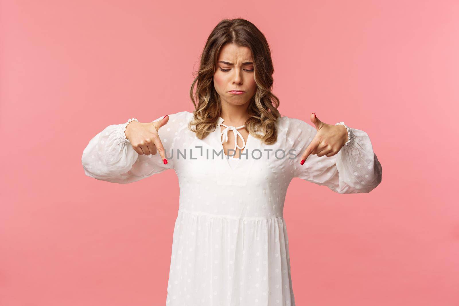 Gloomy, jealous and upset cute blond european woman in white trendy spring dress, sulking as pointing and looking down, see something expensive, cant efford it, strand pink background.