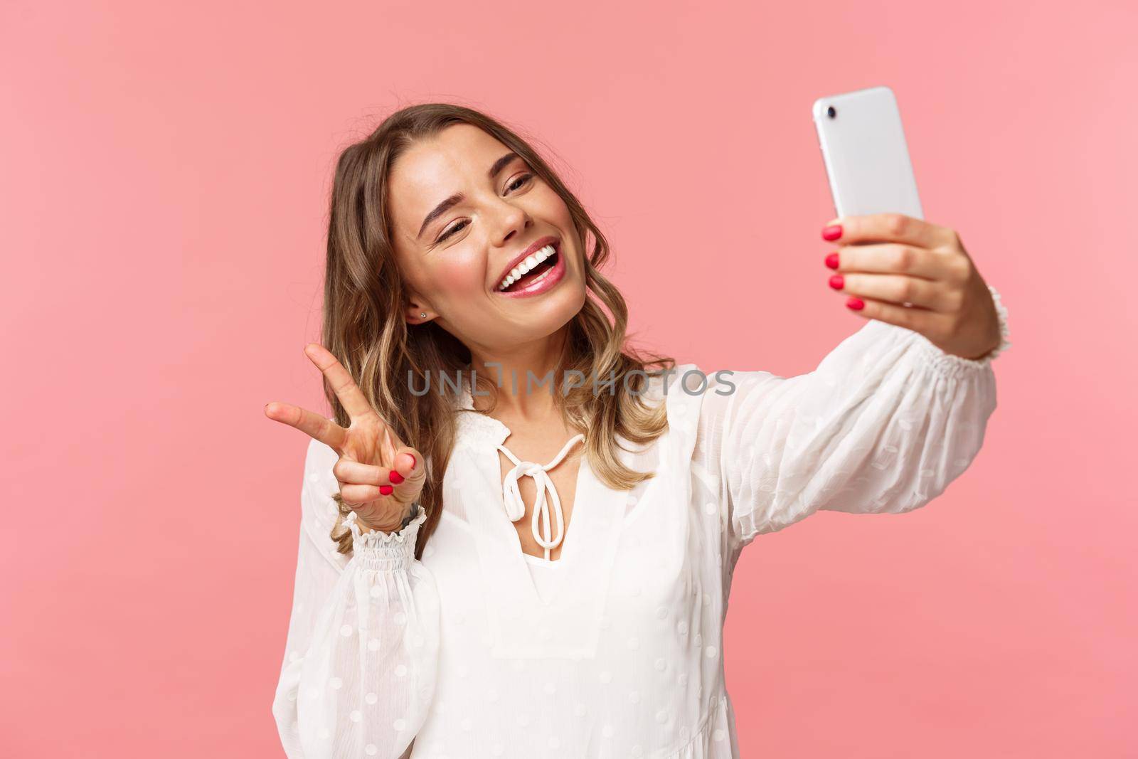 Close-up portrait of cheerful lovely, feminine blond girl in white dress, taking selfie on mobile phone, make kawaii peace sign while take photo, capturing spring moment, pink background.