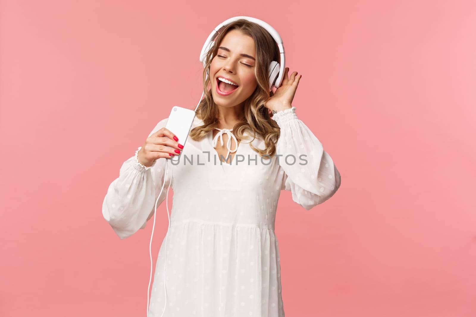 Portrait of beautiful blond caucasian female in white dress, listening music in headphones, close eyes and smiling, using mobile phone like microphone, singing along favorite song, pink background.