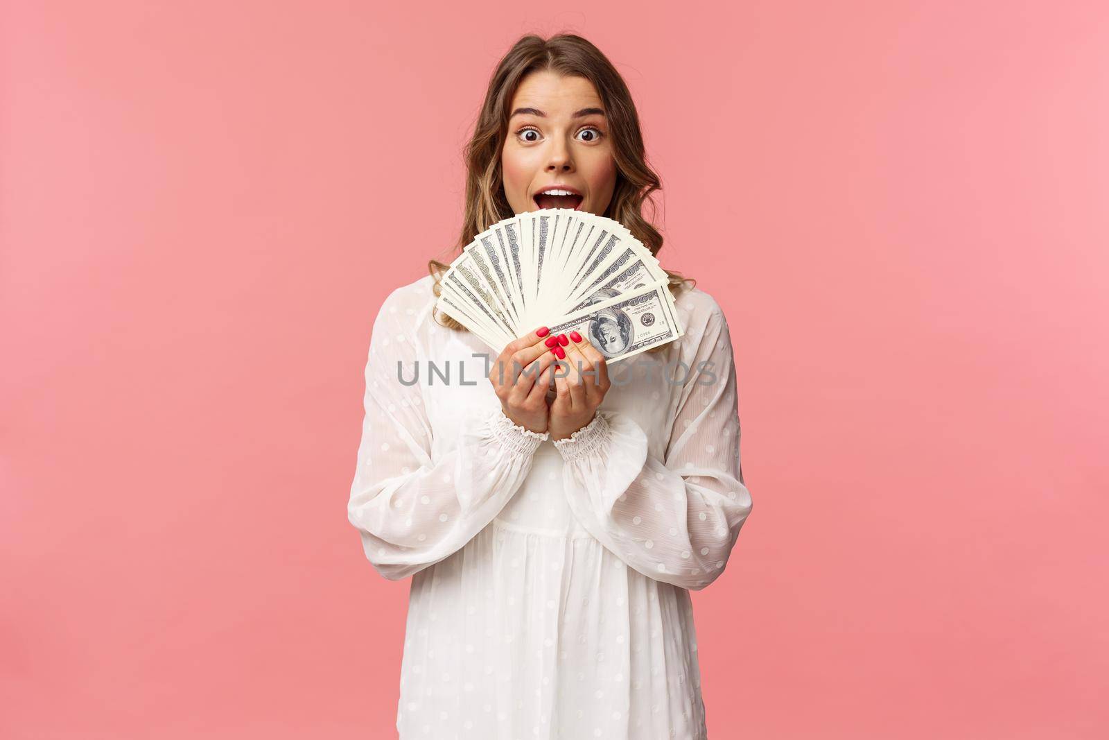 Portrait of excited lucky girl with blond hair, white dress, winning money, receive cash award, big lottery prize, holding dollars near face, smiling and looking amazed, standing pink background.