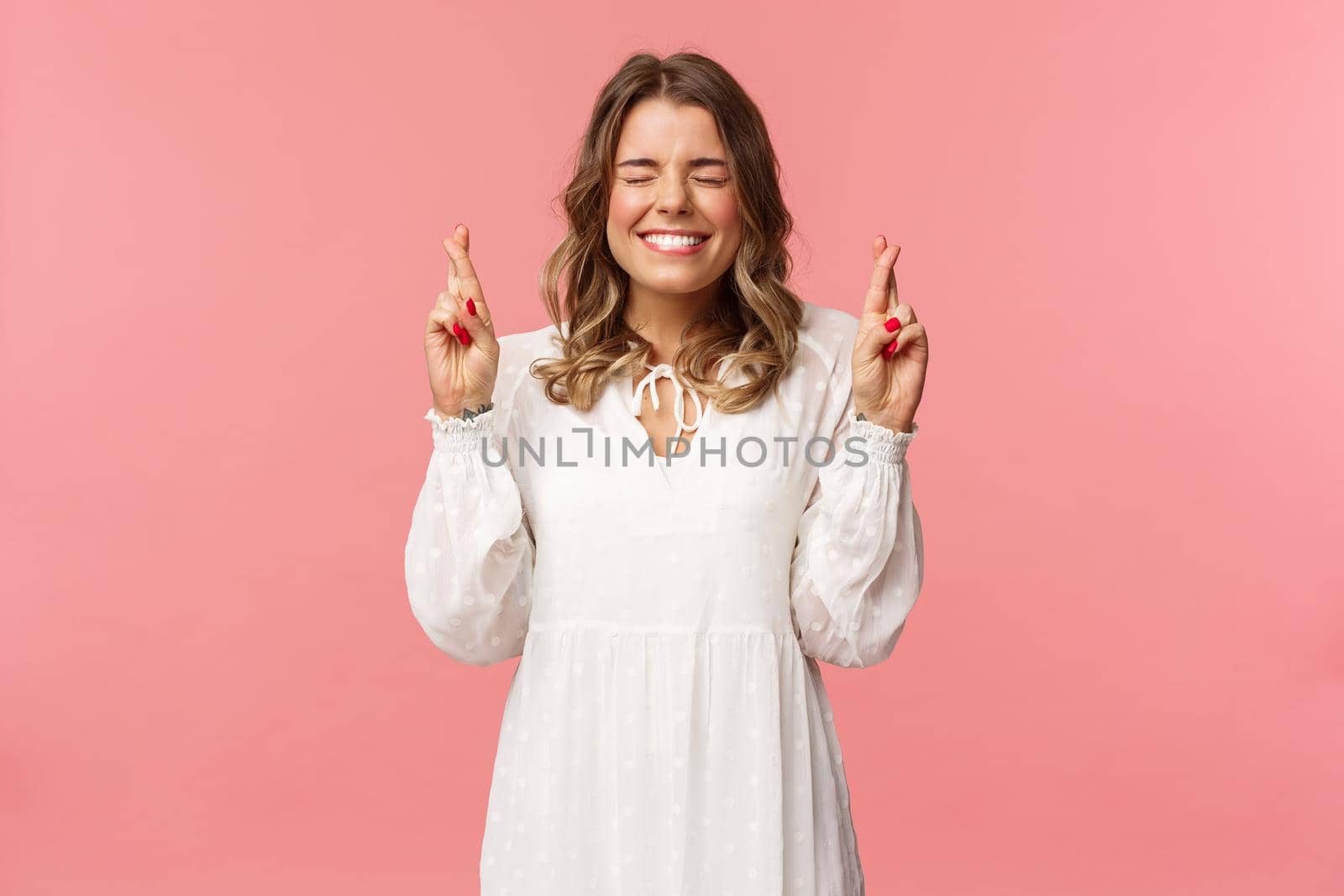 Portrait of hopeful lucky cute blond girl in white dress praying, anticiapte miracle, making wish to win, cross fingers good luck, smiling and close eyes and pleading god, pink background.
