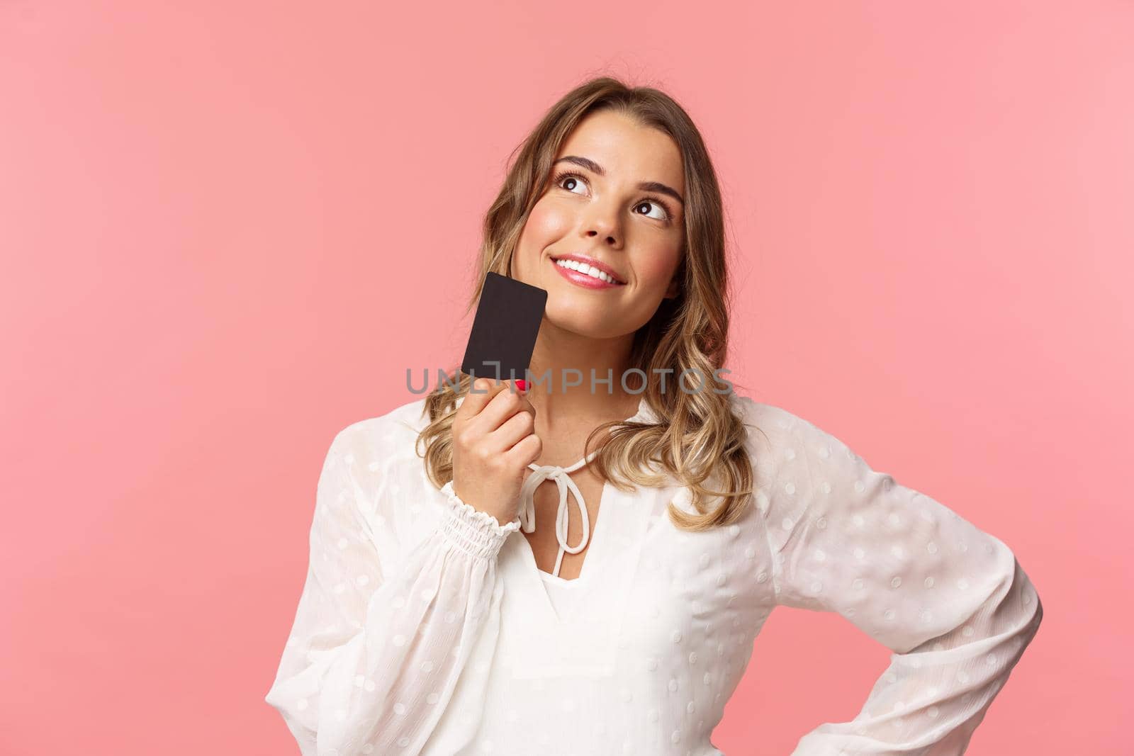 Close-up portrait of thoughtful and creative blond woman in white dress, touching chin with credit card and smiling dreamy as looking up and thinking, picturing what buy as present, pink background.