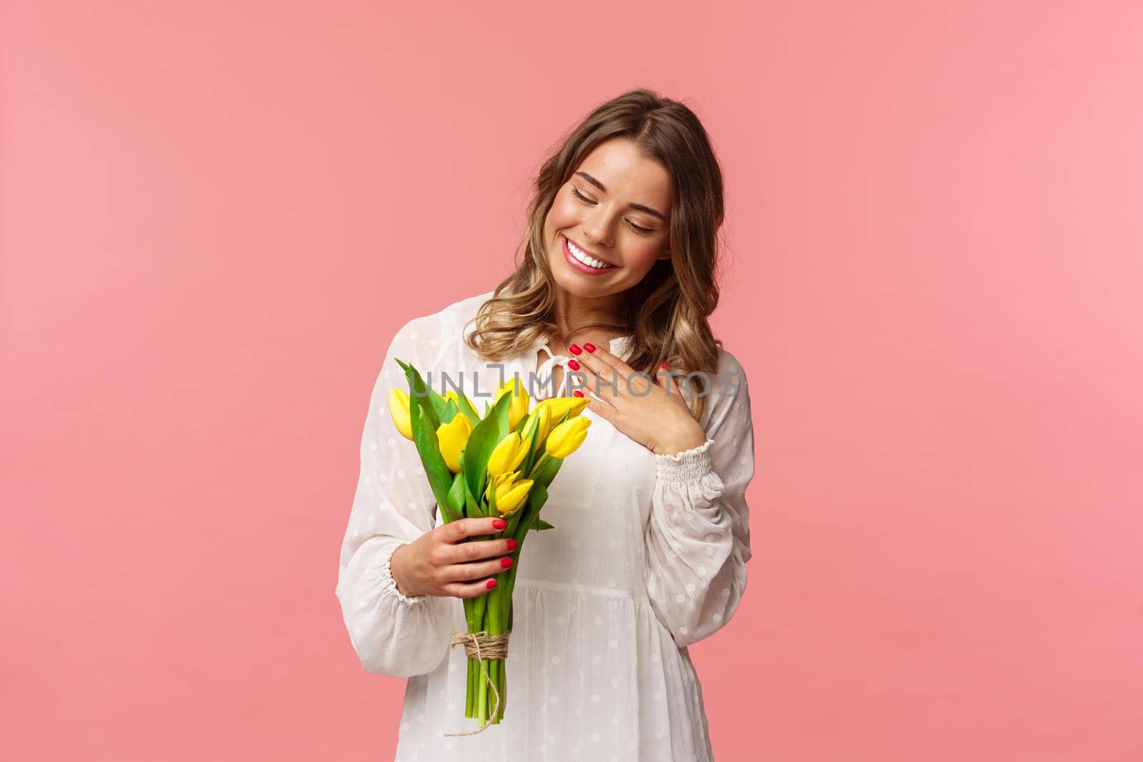 Holidays, beauty and spring concept. Portrait of pleased, cute romantic blond girl receive beautiful bouquet of flowers, looking at yellow tulips and touch heart grateful, smiling.