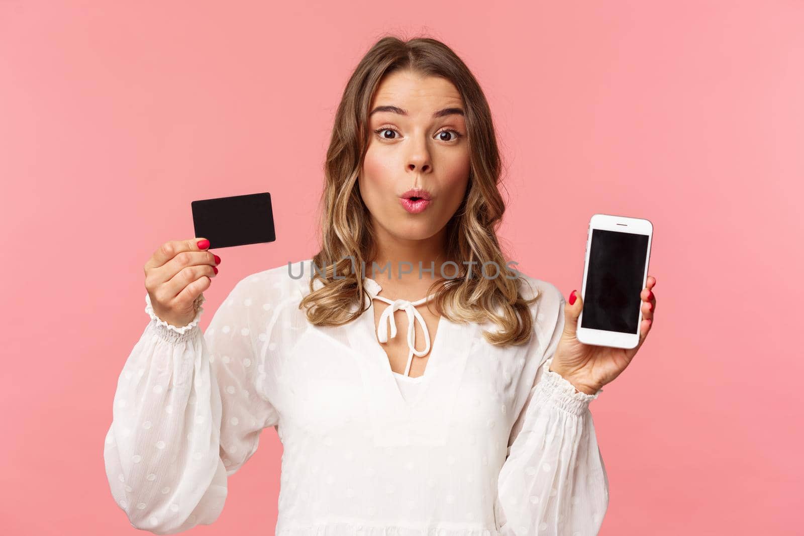 Finance, shopping and technology concept. Close-up portrait of excited blond cute girl in white dress, fold lips amused look camera as showing credit card and mobile phone, pink background by Benzoix