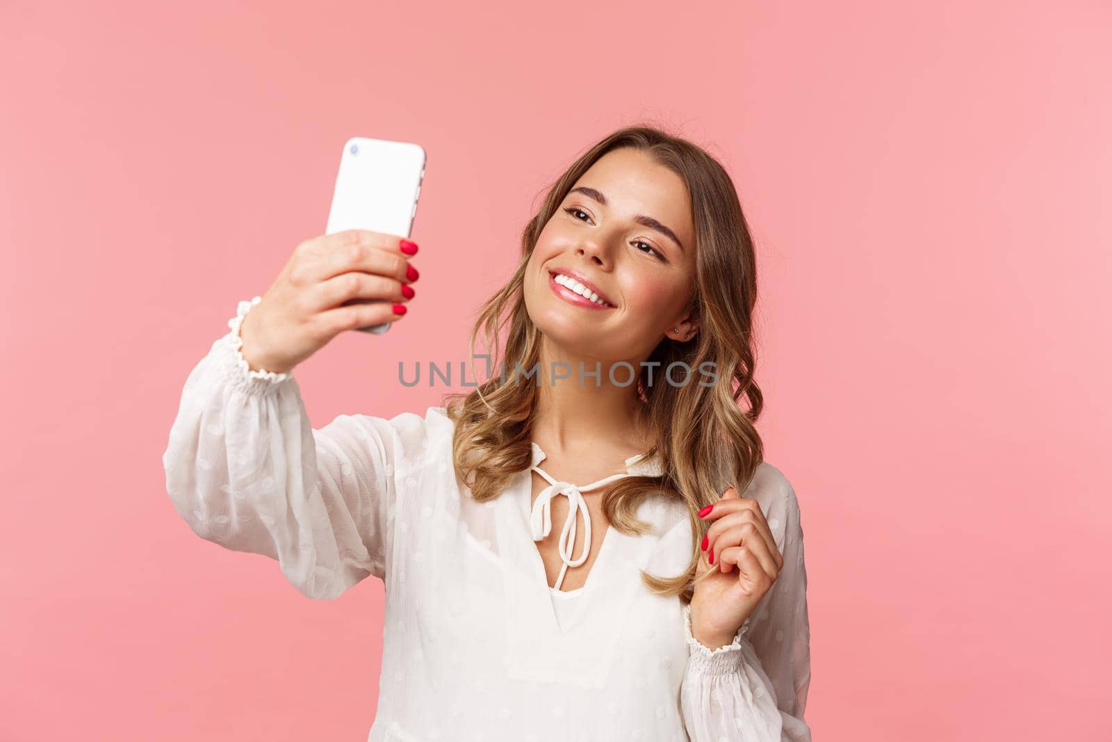 Close-up portrait of beautiful and stylish blond girl taking selfie in park at spring, gather likes and followers, make blogger content, taking photo on smartphone, smiling sassy, pink background.