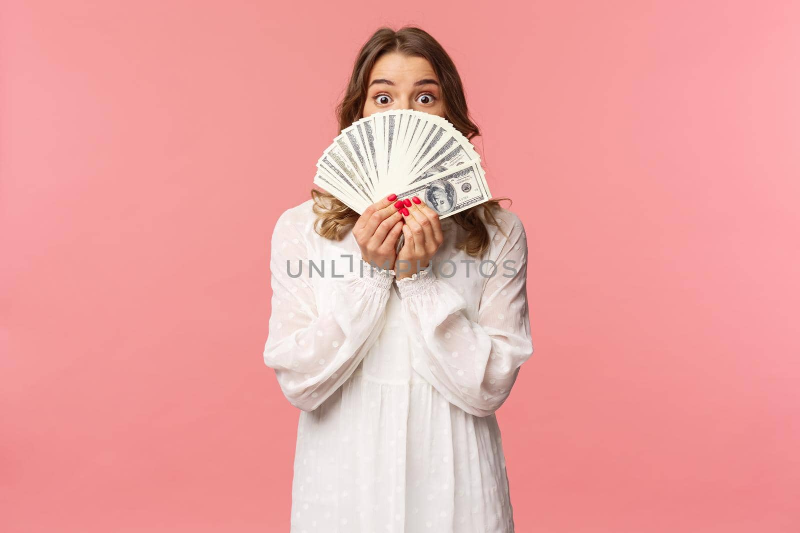 Portrait of amazed and excited cute feminine blond girl in white dress, holding dollars over face, looking from under cash at camera with surprised expression, stand pink background by Benzoix