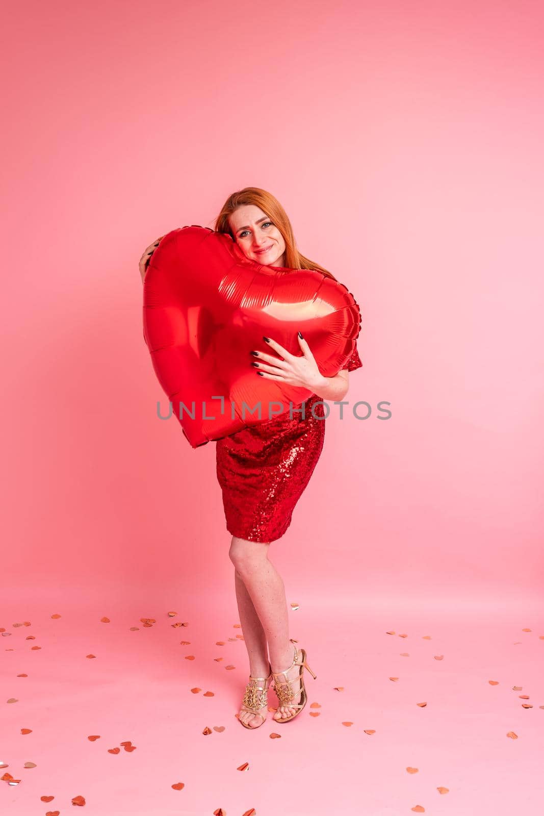 Beautiful redhead girl with red heart baloon posing. Happy Valentine's Day concept. Studio photo of beautiful ginger girl dancing on pink background.