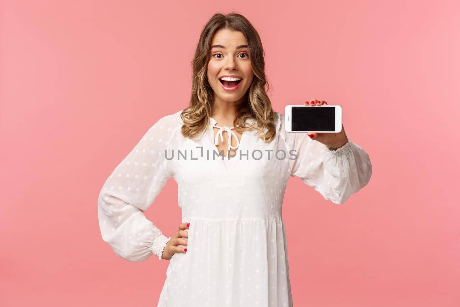 Portrait of cheerful, upbeat attractive blond caucasian girl in white dress, showing smartphone display, hold mobile phone horizontal smiling camera amazed, stand pink background.