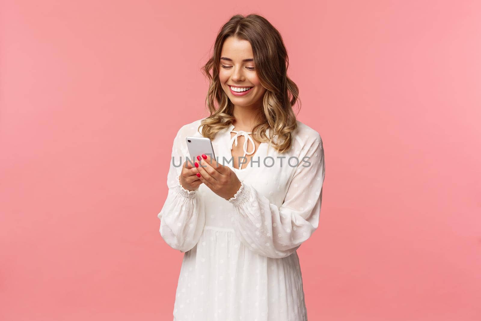 Technology and smartphones concept. Attractive young woman in cute white dress messaging, arrange romantic date, booking tickets via online application on mobile phone, smiling at display by Benzoix